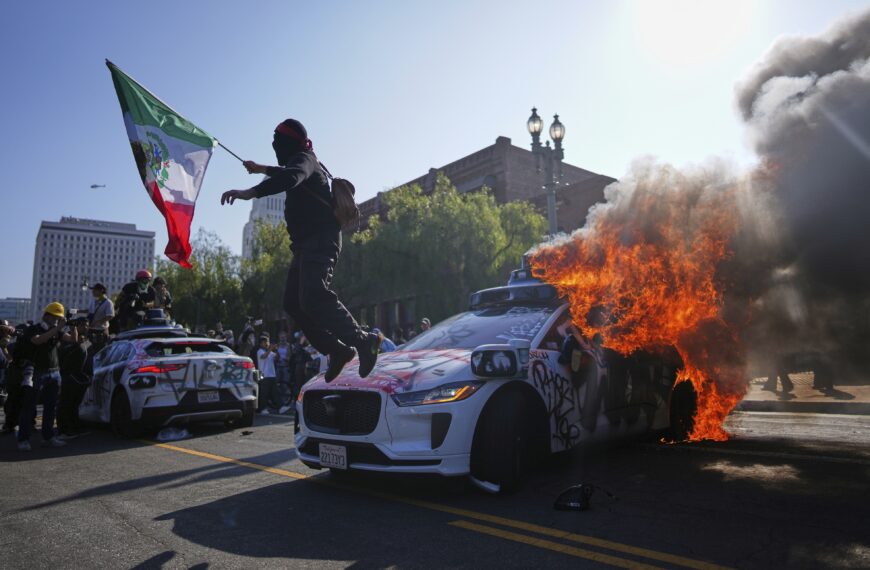 A protester leaps off a burning Waymo taxi near the Metropolitan Detention Center in downtown Los Angeles, Sunday, June 8, 2025, following last night's immigration raid protest. (AP Photo/Eric Thayer)