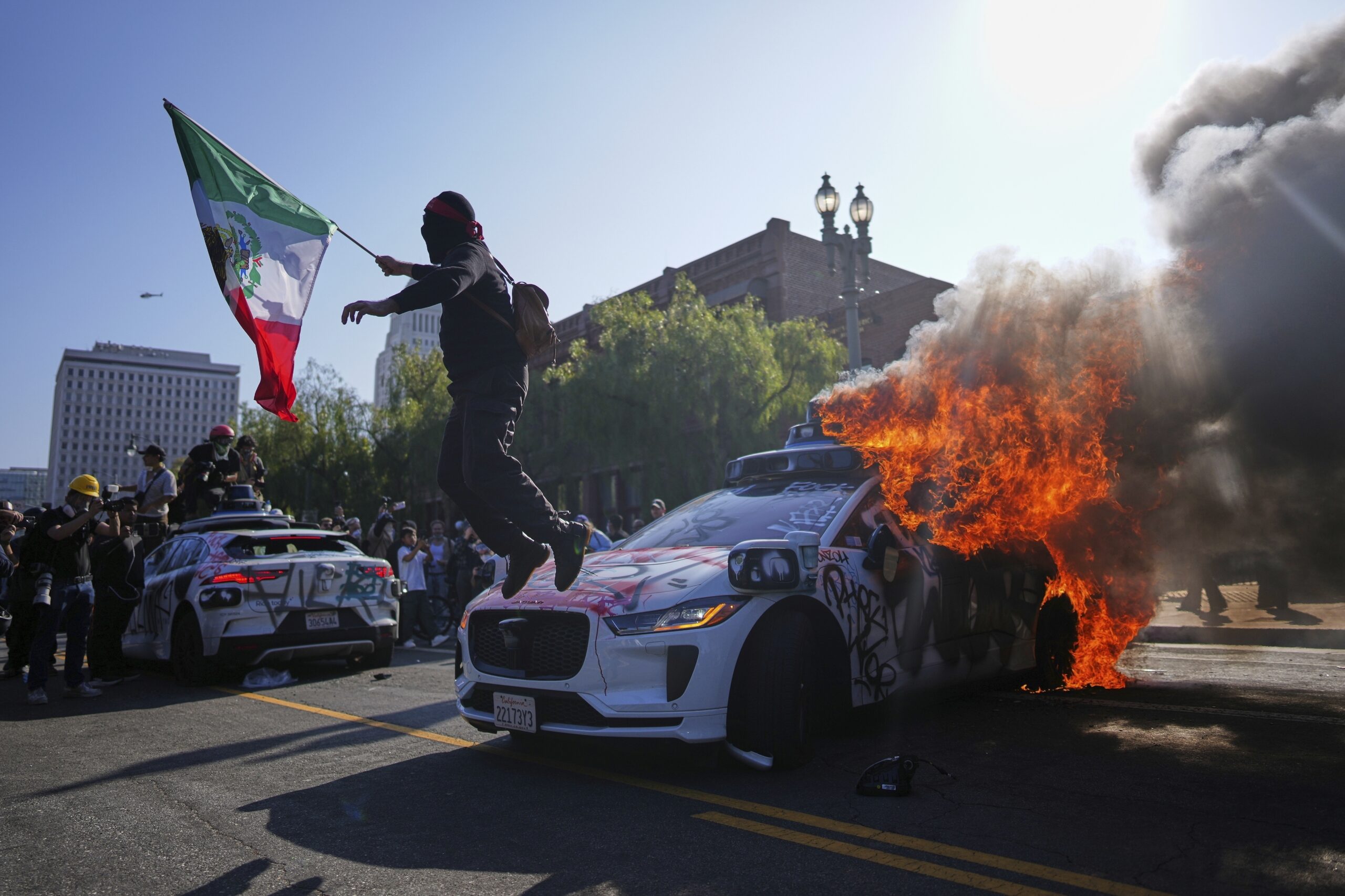 A protester leaps off a burning Waymo taxi near the Metropolitan Detention Center in downtown Los Angeles, Sunday, June 8, 2025, following last night's immigration raid protest. (AP Photo/Eric Thayer)